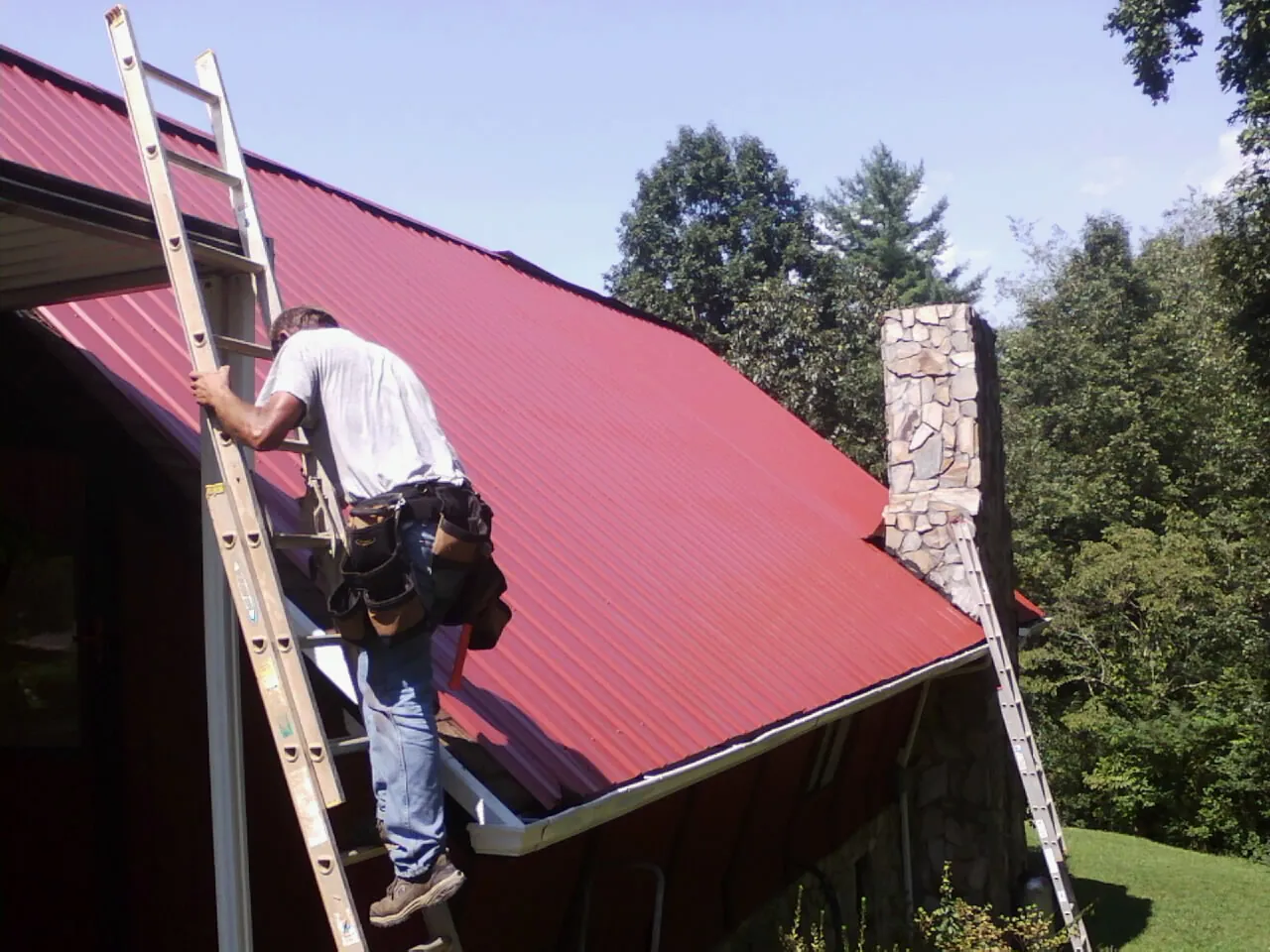 Roofer on a ladder beside a red metal roof and stone chimney — Soffit & Fascia Repair by Roofing Brothers in Sumatra